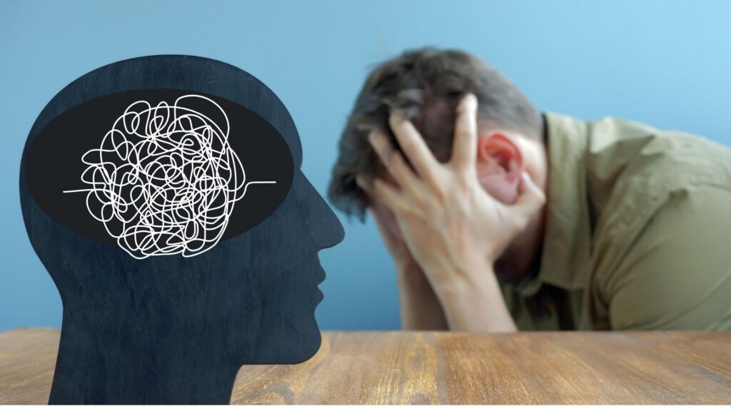 A man is holding his head, depressed and broken. He is in a bad mental state. In the foreground there is a drawing of a head with chaos in the brain.
