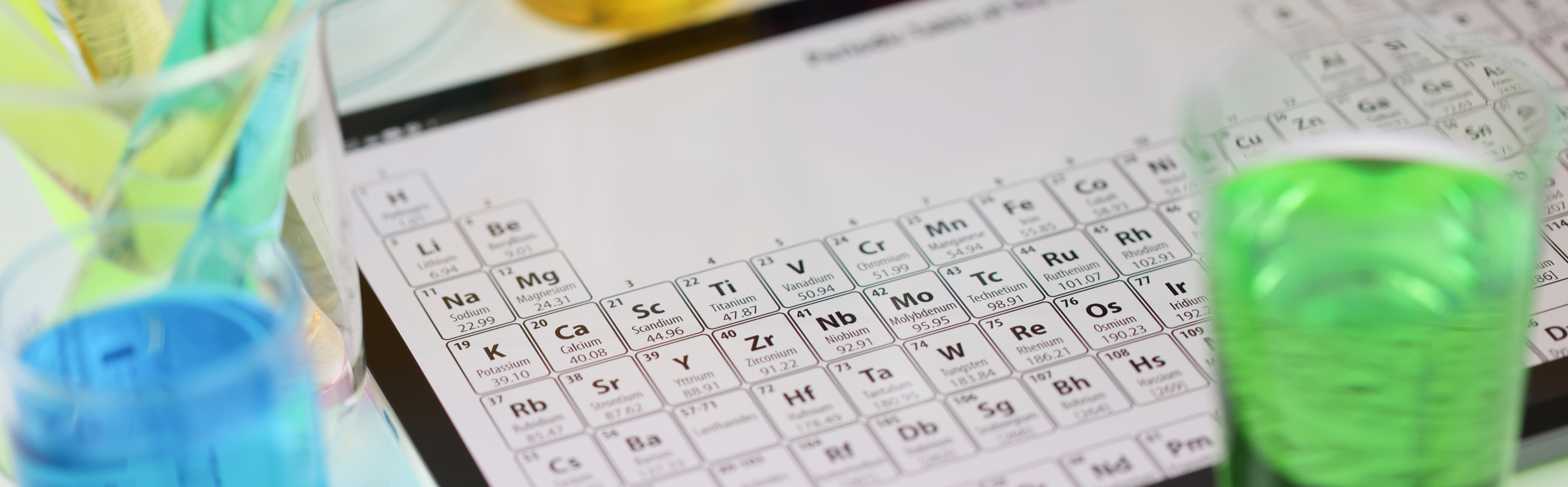 Periodic table of elements in the foreground with laboratory flasks and equipment from a pharmaceutical-chemical lab in the background, representing the analysis and control of elemental impurities (EI) in medicinal products according to ICH Q3D guidelines. The image underscores the importance of assessing and managing EI levels to ensure product safety and regulatory compliance.